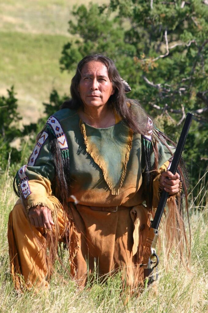 Thomas white crouchin on the ground holding a rifel and waering tradisional native american clothing